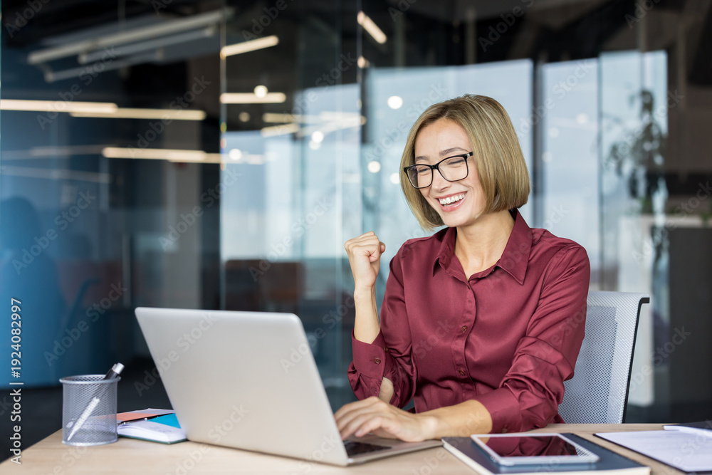 Obraz premium Young businesswoman triumphing on laptop, expressing happiness and excitement after achieving a significant success or completing a project in a modern office environment