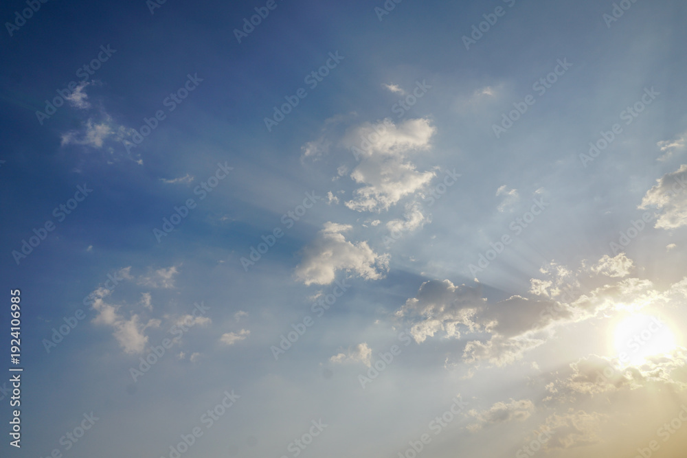 Obraz premium Bright blue sky filled with scattered white clouds and sunlight shining through the clouds. The sun is low enough to emit visible rays in the blue and white sky.