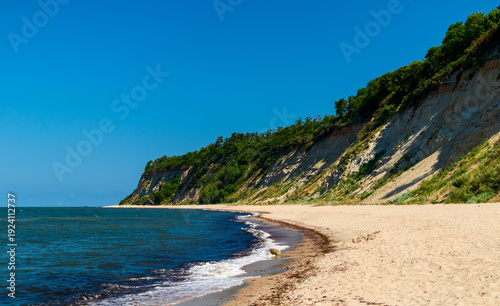 A sandy beach leads the eye toward Cape Taran, where a steep vegetated cliff meets the Baltic Sea. Peaceful coastal scene on a sunny day with blue sky
