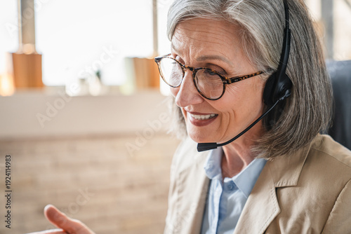 Senior female call center operator talking to customer, using headset with microphone and monitor at workplace, speaking at desktop, giving consultation, information technology support