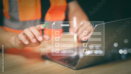 Professional engineer in safety vest using a futuristic digital checklist interface over laptop for quality control, project management, and construction site safety audit process.