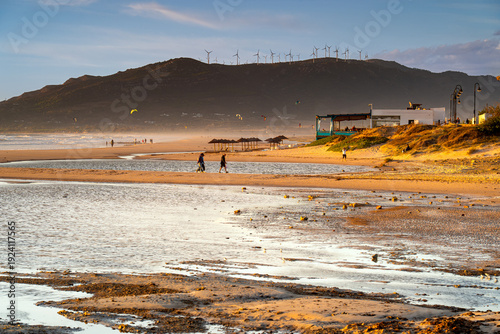 People walk along the beach at sunset with kites flying in background, Tarifa, Spain.