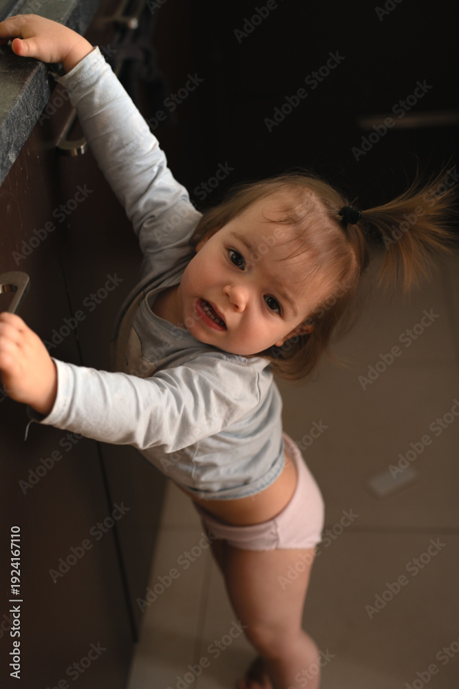 Fototapeta premium curious toddler reaching and leaning on a dark kitchen cabinet countertop in soft warm light, candid domestic portrait highlighting skin tones, muted browns and pinks, playful intimate expression