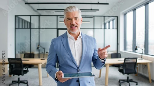 Mature businessman holding a tablet in a modern office setting with a surprised expression on his face