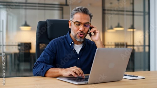 Man sitting at desk working on laptop and talking on phone in office