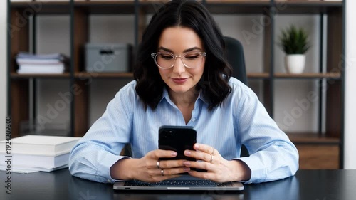 Woman sitting at office desk looking at smartphone and laptop