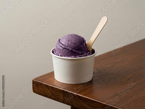 A cup of taro or ube gelato with a wooden ice cream spoon, placed on the edge of a brown table, suitable for food advertisement or food menu 