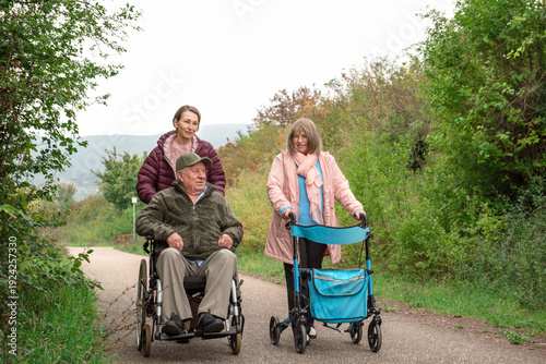 Happy family taking a walk together with daughter pushing her father in wheelchair and mother walking with help of walker, smiling happy and talking