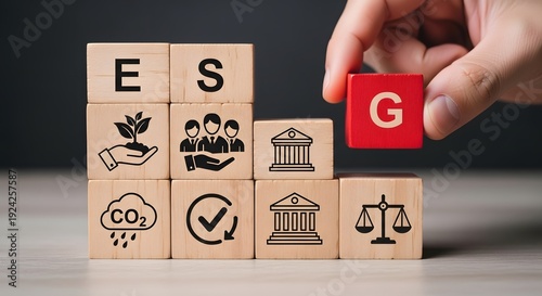 A hand holding a red block with a white letter g above wooden blocks with esg icons on a table