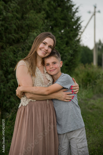 A young Caucasian woman with long brown hair embraces a boy with short brown hair in a green outdoor setting. They both smile happily.