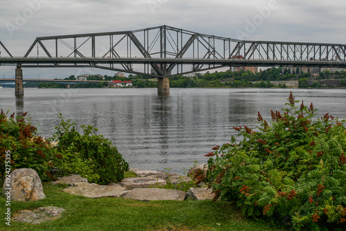  Ottawa, Ontario, Canada. View of the wide steel truss Alexandra bridge spanning the Ottawa river, with green shrubs and rocks in the foreground.
