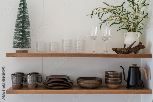 Kitchen open shelf with ceramic plates, mugs and bowls, drinking glasses and a plant. 