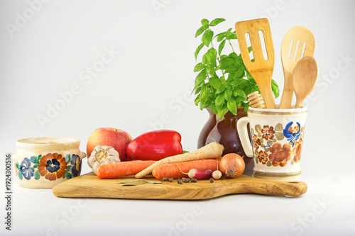Traditional Polish ceramic dishes with fresh vegetables, wooden kitchen utensils, and potted basil on a white background.