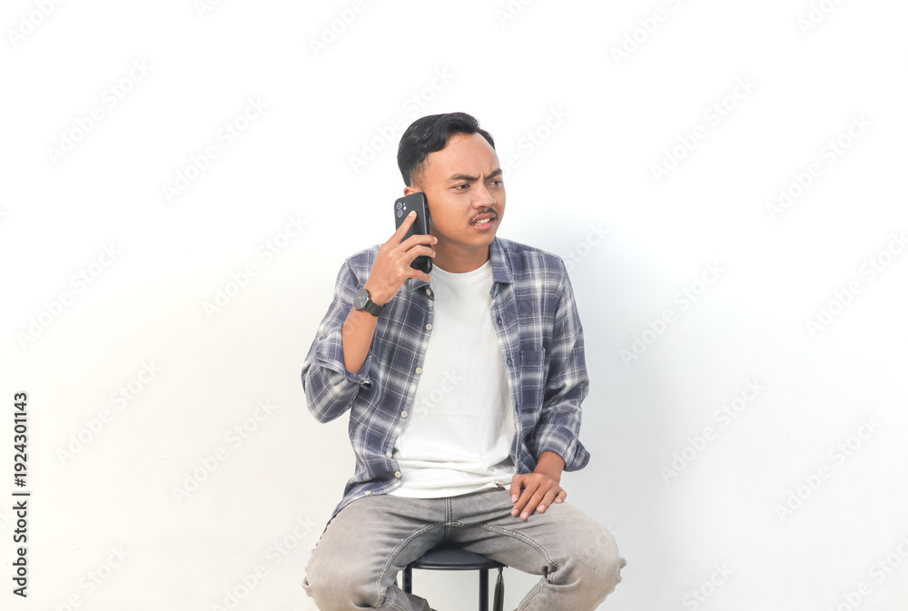 Obraz premium Asian man wearing a plaid shirt with a confused expression is talking on the phone on an isolated white background.