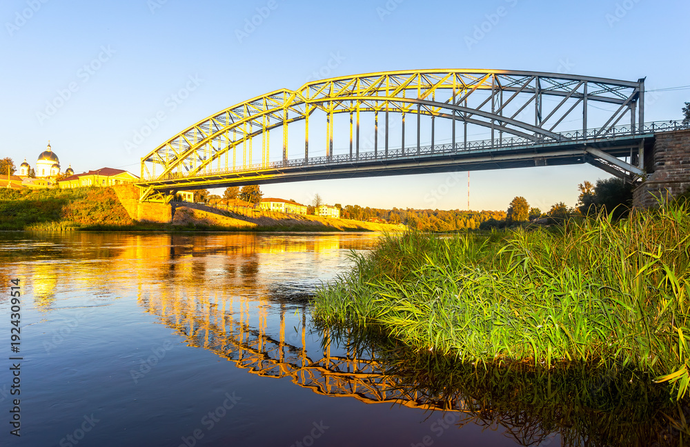 Obraz premium View on the steel arch bridge across the Msta river in summer in sunset light. Built in 1905. Borovichi, Russia