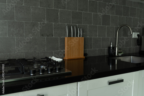Modern kitchen interior featuring a black countertop, stainless steel sink, gas stove, and wooden knife block against a textured gray tile backsplash