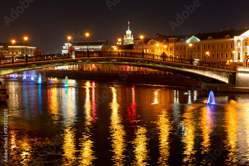 Bright lights reflect on the water in Moscow at night near a bridge