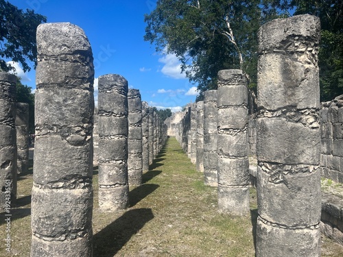 View down rows of grey stone pillars, Mayan ruins, green vegetation, blue skies, November, at Chichen Itza, Yucatan, Mexico
