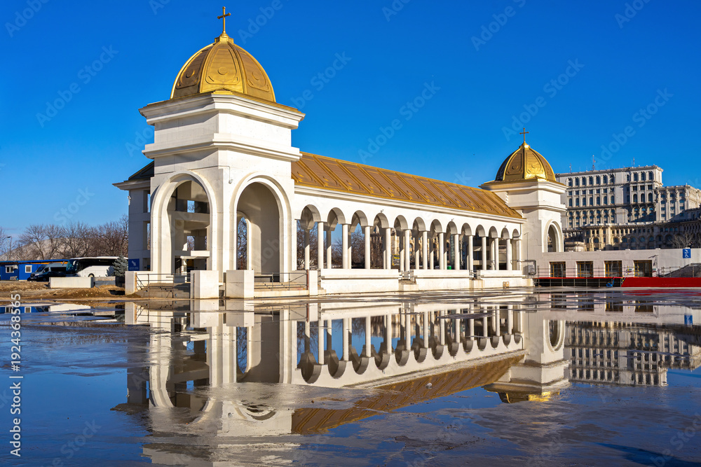 Naklejka premium Colonnaded pavilion with golden domes near the Romanian National Cathedral in Bucharest, reflected in rainwater under a clear blue sky in the capital of Romania.