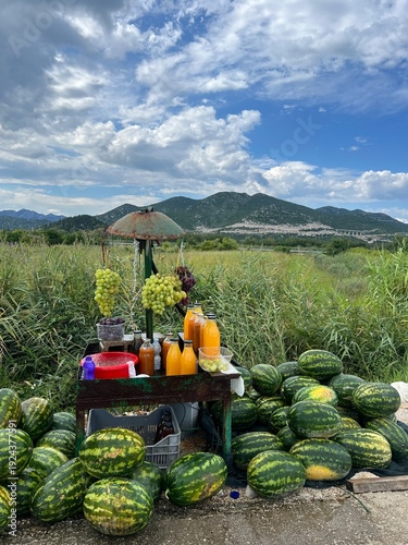 Roadside fruit stand with watermelons, grapes and juice, green grass and vegetation in back, mountains in distance, cloudy skies, north of Dubrovnik, Croatia