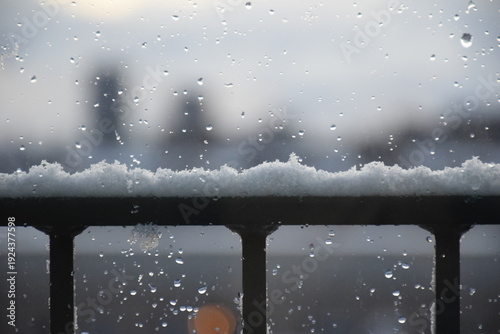 Close-up view of white snow on horizontal metal railing, drops on window, blurry buildings in back, grey skies, winter time, Copenhagen, Denmark