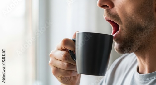 Man yawning while holding dark grey mug, enjoying his morning coffee near window. Morning coffee ritual provides energy, awakening mind and body for day ahead.