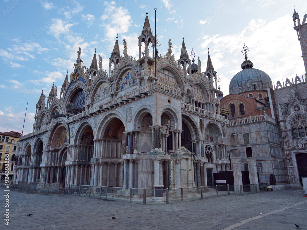 Fototapeta premium St Mark's Basilica Facade and Mosaics - Wide, majestic view of the ornate Byzantine facade of St. Mark's Basilica with marble arches, detailed mosaics, and spires under a cloudy blue sky.