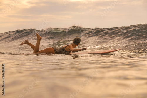 Konstfotografi Young Woman Paddling On Surfboard At Sunset, Lying Prone And Stroking Toward Hor