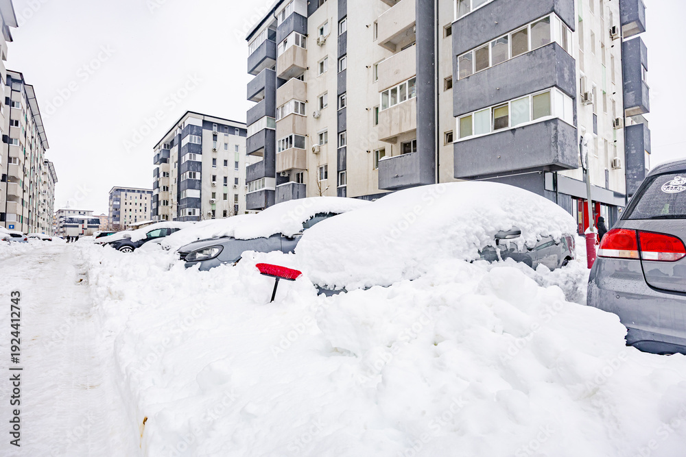 Fototapeta premium Red snow brush sticking out of deep snow covering parked cars in residential courtyard after heavy winter blizzard in the city