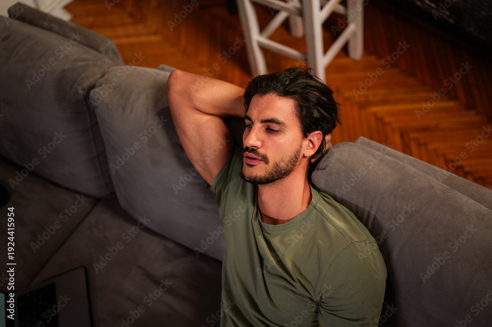 Obraz premium Young man wearing a green t shirt relaxing on a comfortable gray couch, contemplating or experiencing exhaustion, with one arm resting behind his head in a casual pose at home