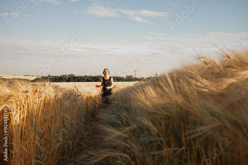 Young woman in black dress walking through golden wheat field at sunset. Cinematic nature landscape, summer freedom and lifestyle concept. Soft bokeh foreground