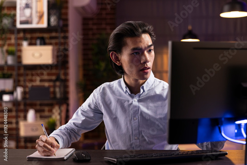 Self employed asian man studying online late at night at home office, using desktop computer for business learning. Focused male individual taking notes while catching up on digital courses.