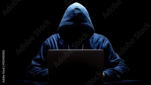 Anonymous hacker in hoodie sits at a laptop on a black background. Cybersecurity concept, cybercrime and digital threat of modern times.