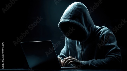 Anonymous hacker in hoodie sits at a laptop on a dark background. Cybersecurity concept, cybercrime and digital threat of modern times.