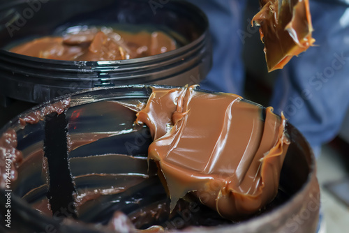 ​A worker uses a spatula to scoop thick brown industrial grease from a black bucket in a workshop, providing lubrication for heavy machinery and automotive parts maintenance.