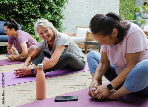 Female friends stretching on purple mats on patio, senior woman reaching feet, coral bottle visible