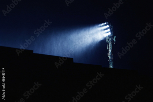 A tall stadium floodlight tower illuminates a dark sports arena at night, with powerful beams of light cutting through mist and rain. The silhouette of a stadium stand creates a dramatic foreground ag