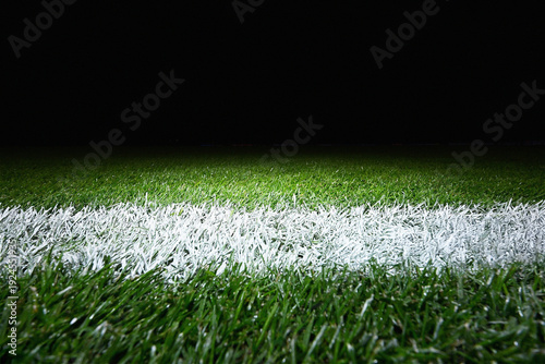 A close-up view of a vibrant green football pitch with a crisp white line, illuminated by stadium lights at night. This low-angle shot captures the detailed texture of the grass and the precision of t