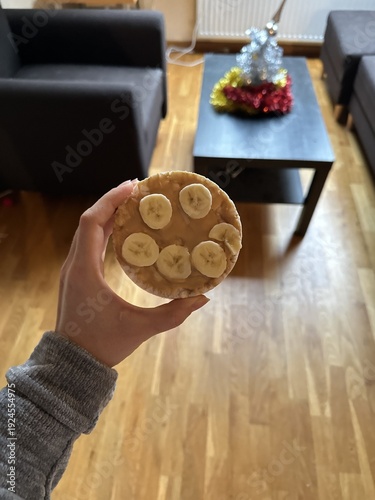 Hand holding a rice cake topped with banana slices in a cozy living room with wooden flooring and a coffee table decorated for the holidays