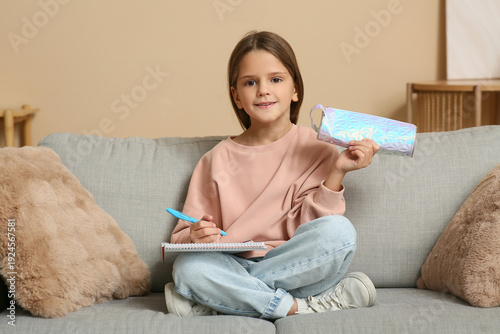 Cute little happy girl with pencil case, pen and notebook sitting on sofa at ...
