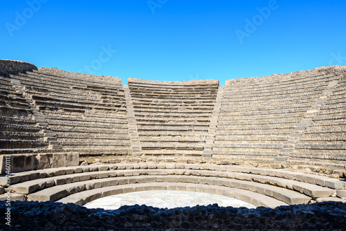 Curved rows of ancient stone seats form the semicircular amphitheater of the small theater in Pompeii, Italy. Bright sunlight highlights the geometric lines of the stepped masonry under a vivid blue