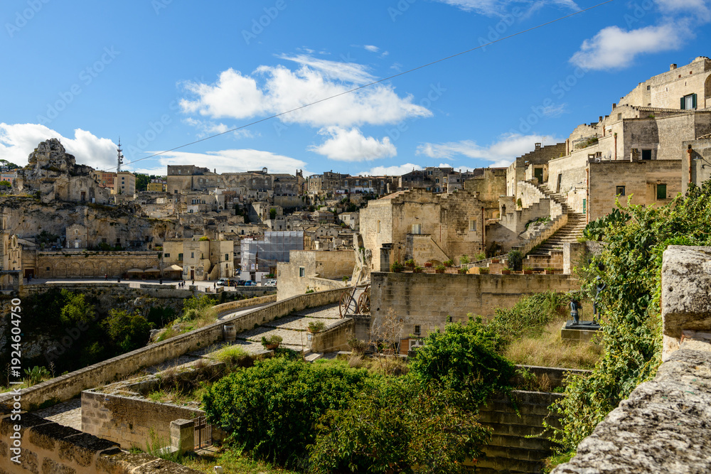 Obraz premium Expansive view of historic stone buildings and terraced rooftops in Matera, Italy, under a clear blue sky with scattered clouds. Sunlight highlights textured limestone architecture and lush greenery
