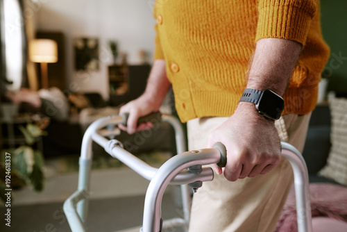 Photos Senior Caucasian man using walker indoors, hands gripping handles while moving f