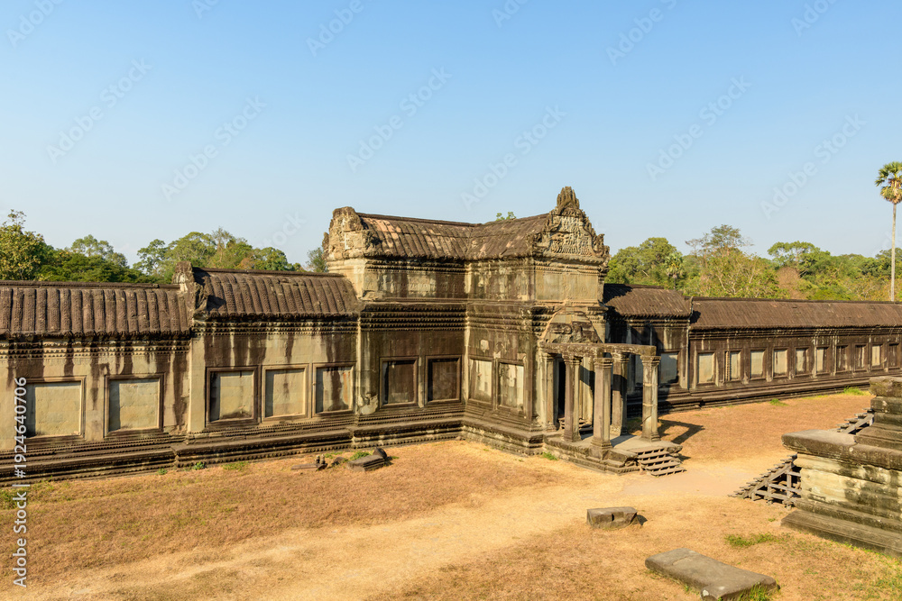 Fototapeta premium Historic sandstone gallery with an ornate entrance pavilion lines the second-level courtyard of Angkor Vat, Cambodia. Weathered stonework and geometric rooflines stand out in the warm afternoon sun