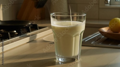 Fresh white milk is gracefully poured from a container into a transparent glass cup, creating a gentle splash on a warm kitchen counter, beautifully illustrating healthy breakfast concepts