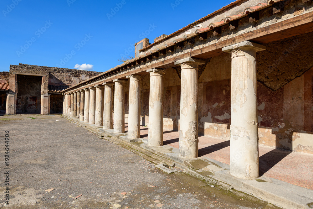 Fototapeta premium Ancient Roman columns line a sunlit portico in the Stabian Baths of Pompeii, Italy. The weathered stone and faded frescoes are highlighted by crisp blue sky, evoking the preserved grandeur of this