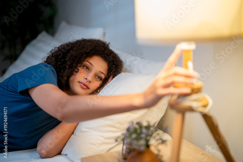 Foto Latino woman lying on bed at night, reaching out to turn off bedside lamp