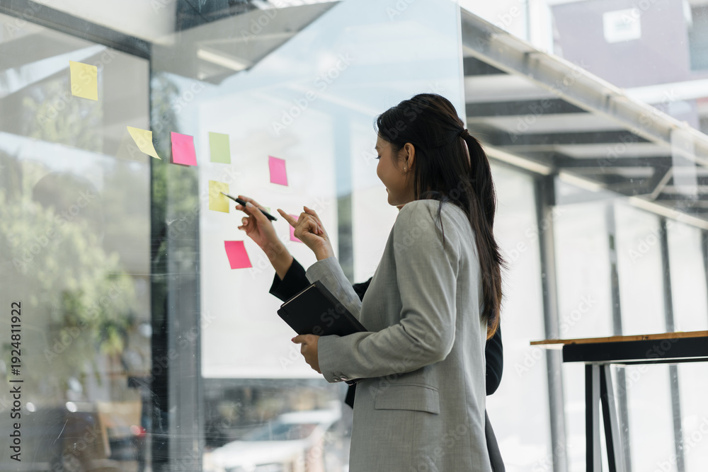 Fototapeta premium Happy Asian businesswoman brainstorming ideas on glass wall with sticky notes while colleague holds tablet in modern office meeting