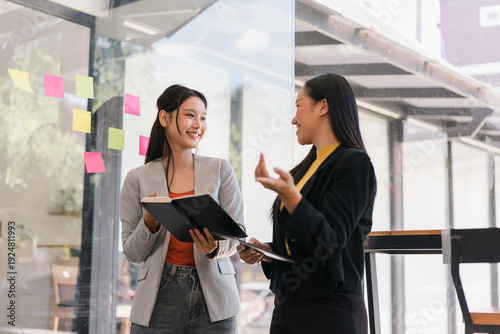 Two happy Asian businesswomen standing talking in modern office holding notebook smiling during corporate meeting discussion