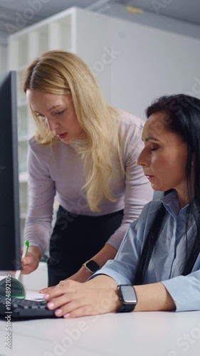Female colleagues collaborating at office workstation, focused woman mentoring coworker on computer task, discussing project details and reviewing information on screen